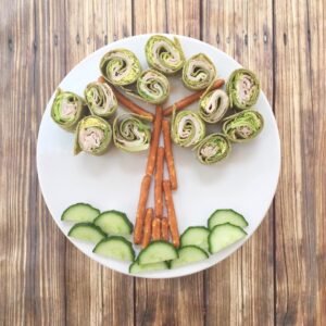 A white plate of cucumber slices, pretzels, and pinwheel sandwiches arranged as a tree